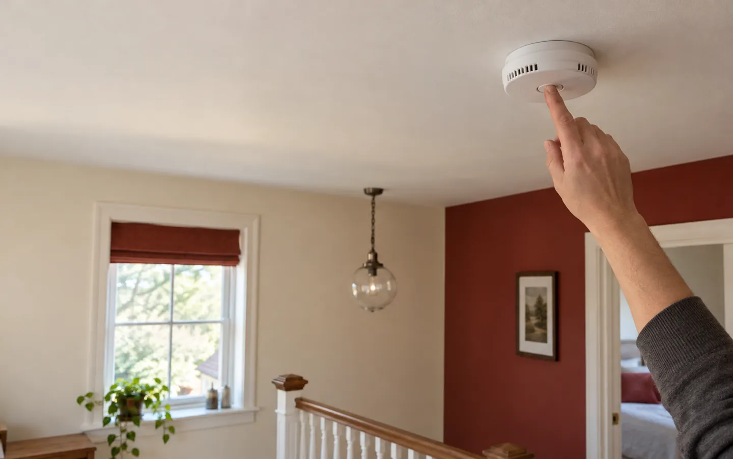 An adult hand reaching up to press the test button on a white smoke alarm on a landing ceiling in soft daylight